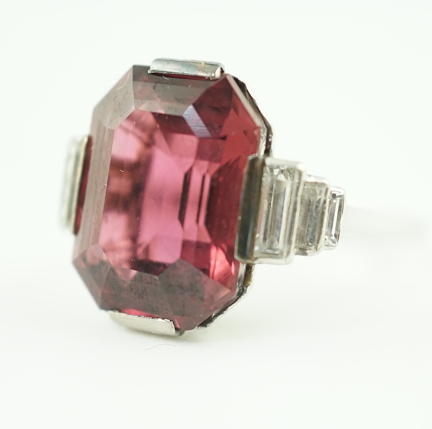 An Art Deco platinum and single stone emerald cut deep pink tourmaline set dress ring, with graduated six stone baguette cut diamond set shoulders